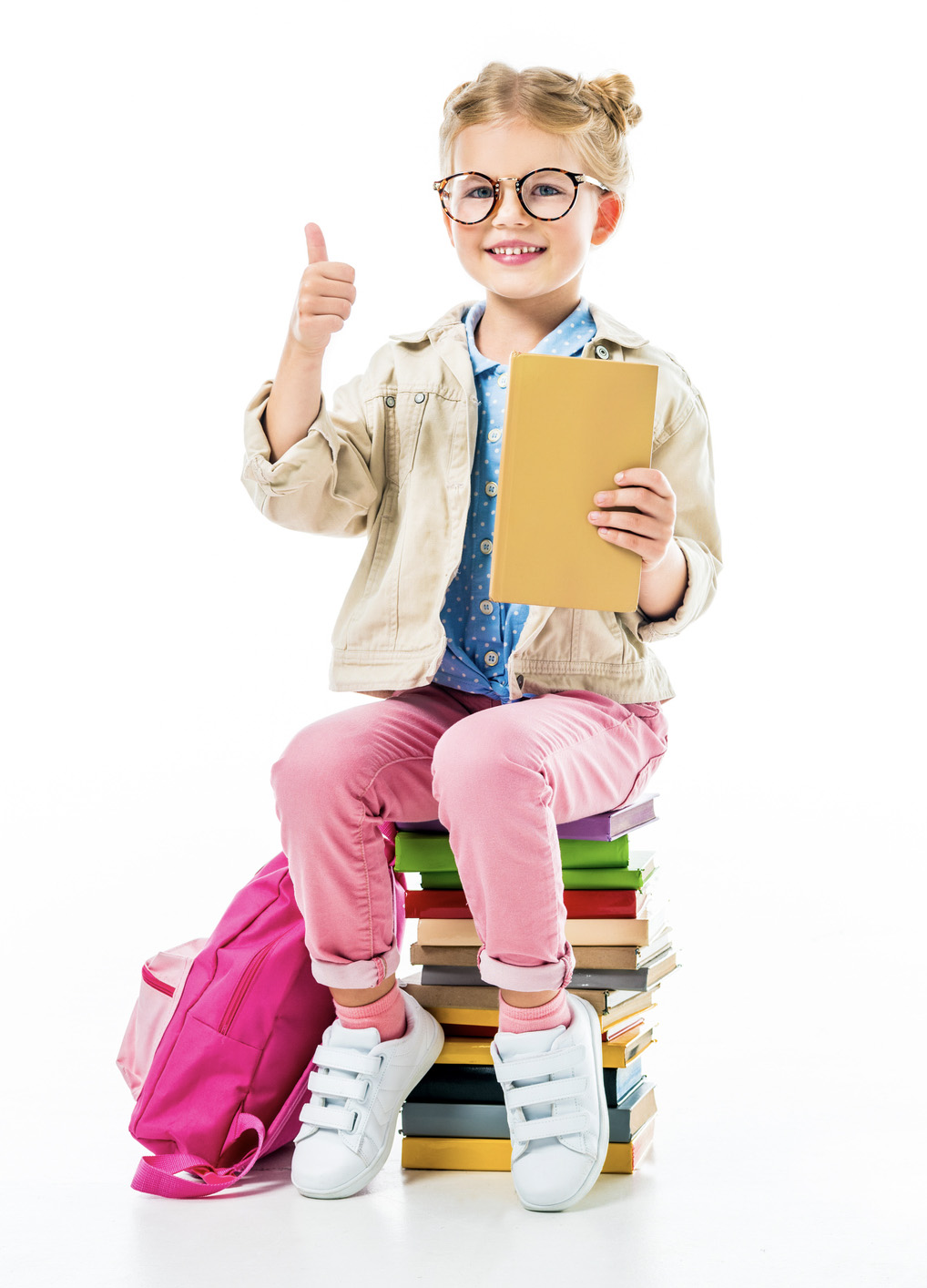 adorable smiling pupil showing thumb up while sitting on pile of books isolated on white, knowledge concept